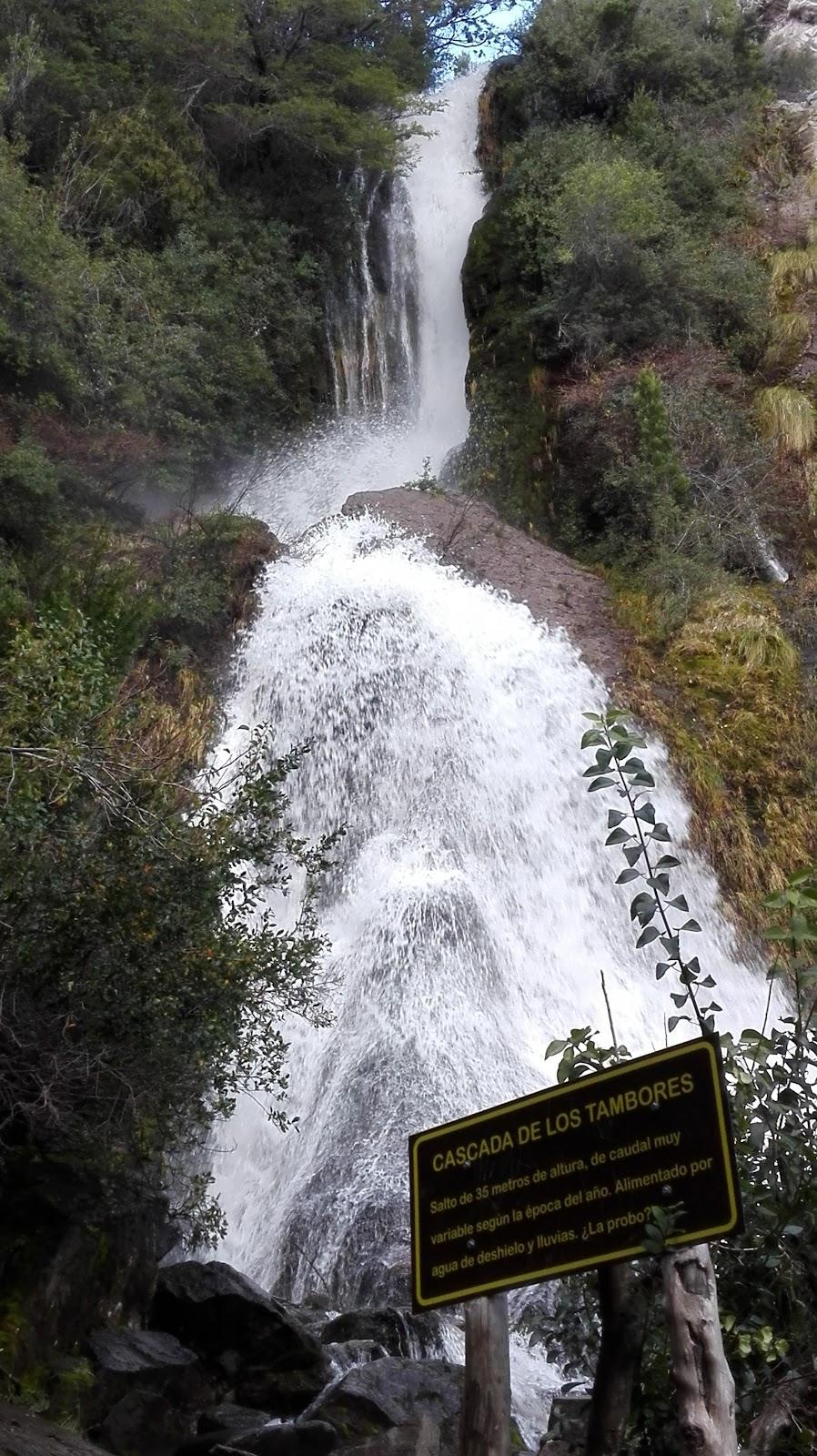 La Cascada de los Tambores cayendo entera entre la vegetación, con el cartel del salto al pie