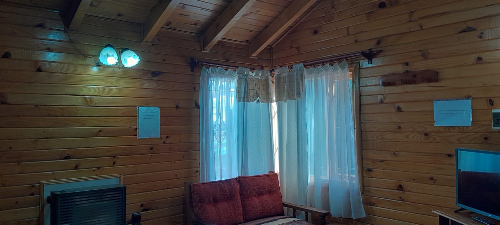 Living de paredes de pino con el sillón, el ventanal con cortinas claras y el televisor